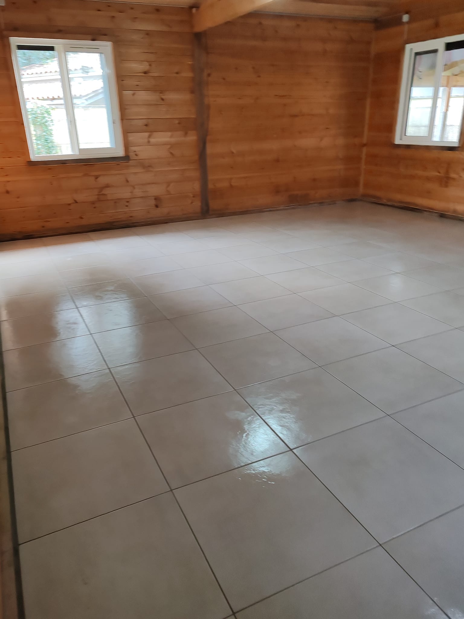 Sol en carrelage gris brillant dans une pièce rustique avec murs en bois et fenêtres blanches.