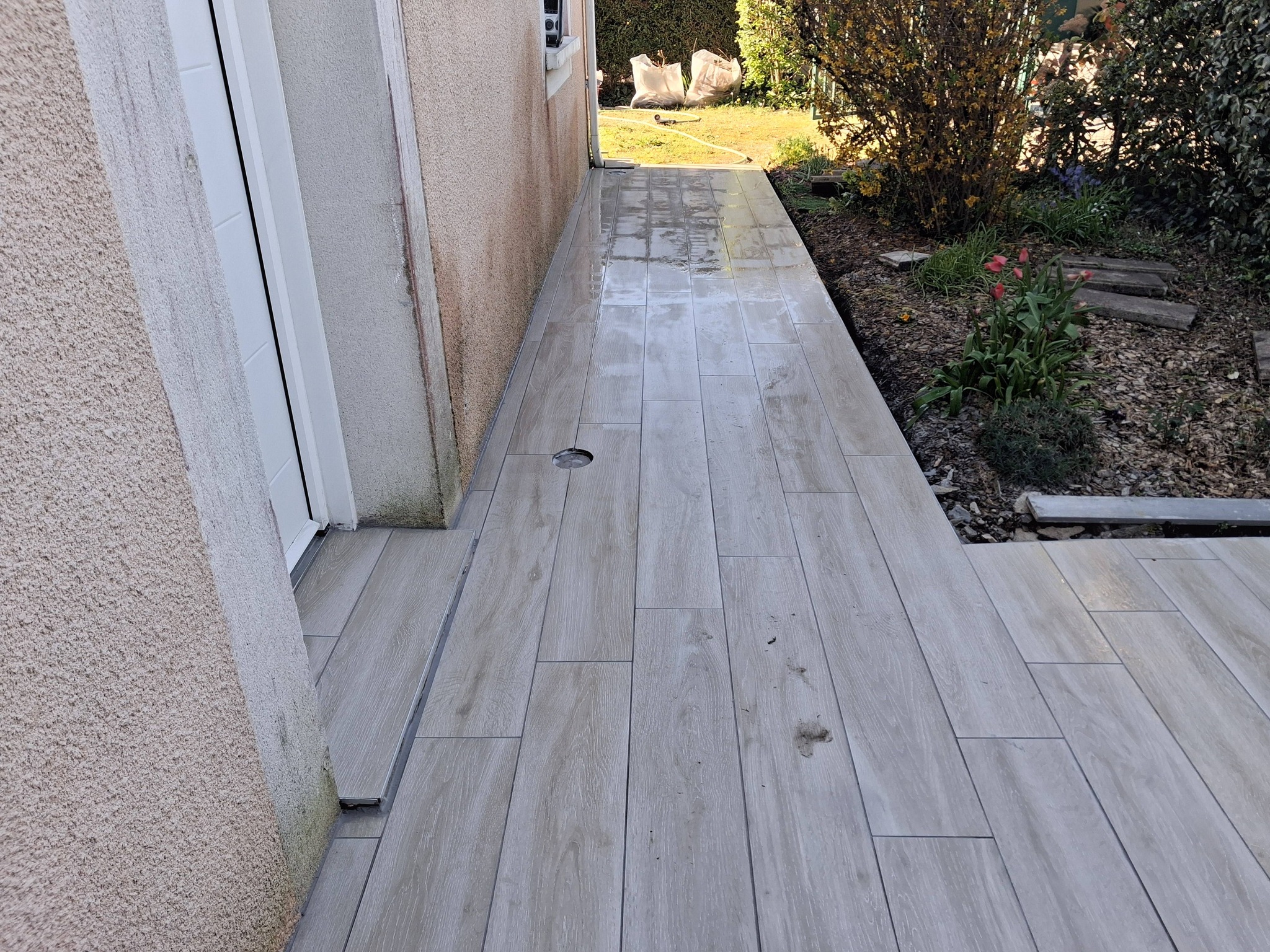 Allée extérieure en carrelage gris aspect bois posé le long d'un mur de maison avec vue sur le jardin.