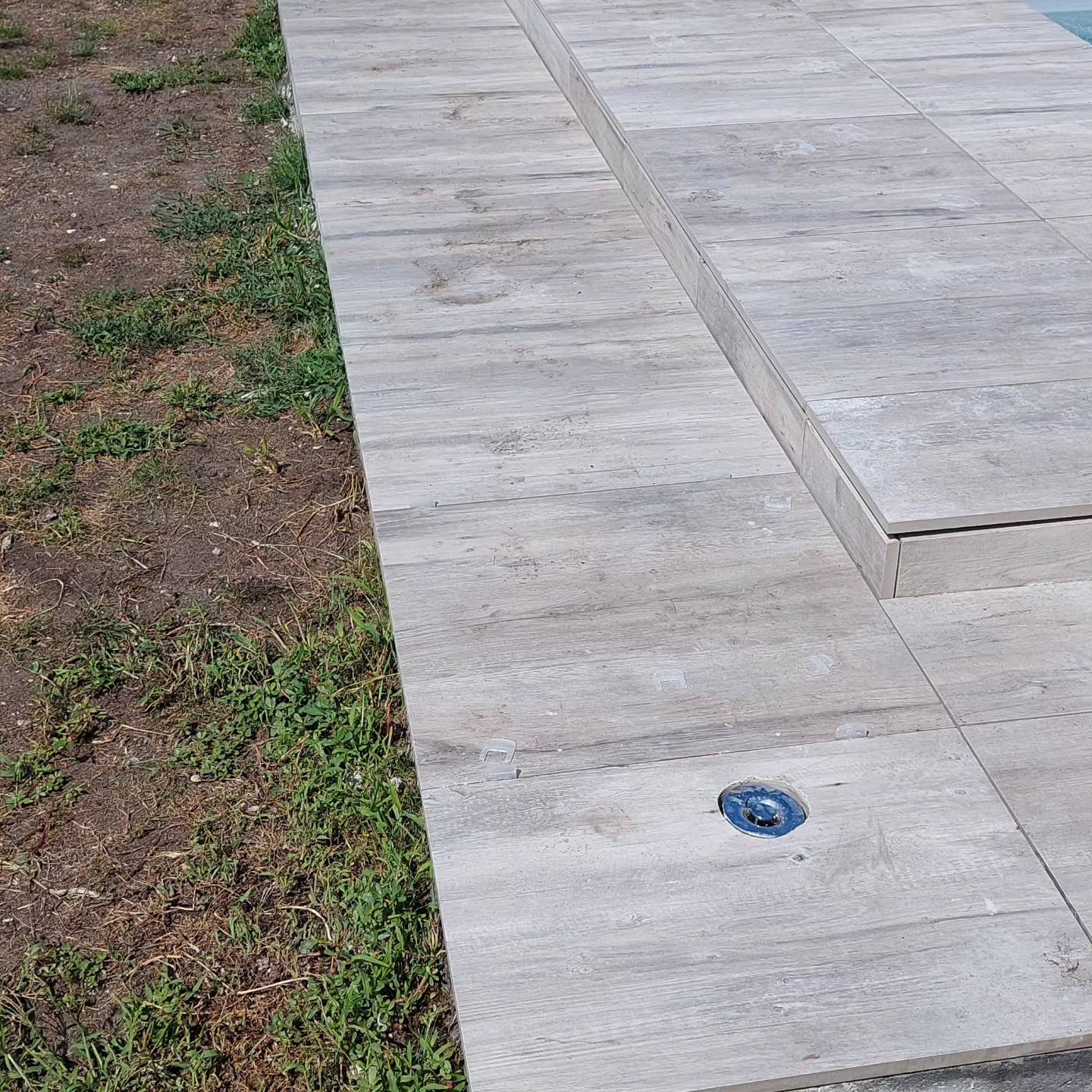 Détail de carrelage extérieur gris aspect bois sur une terrasse de jardin avec une évacuation d'eau bleue.