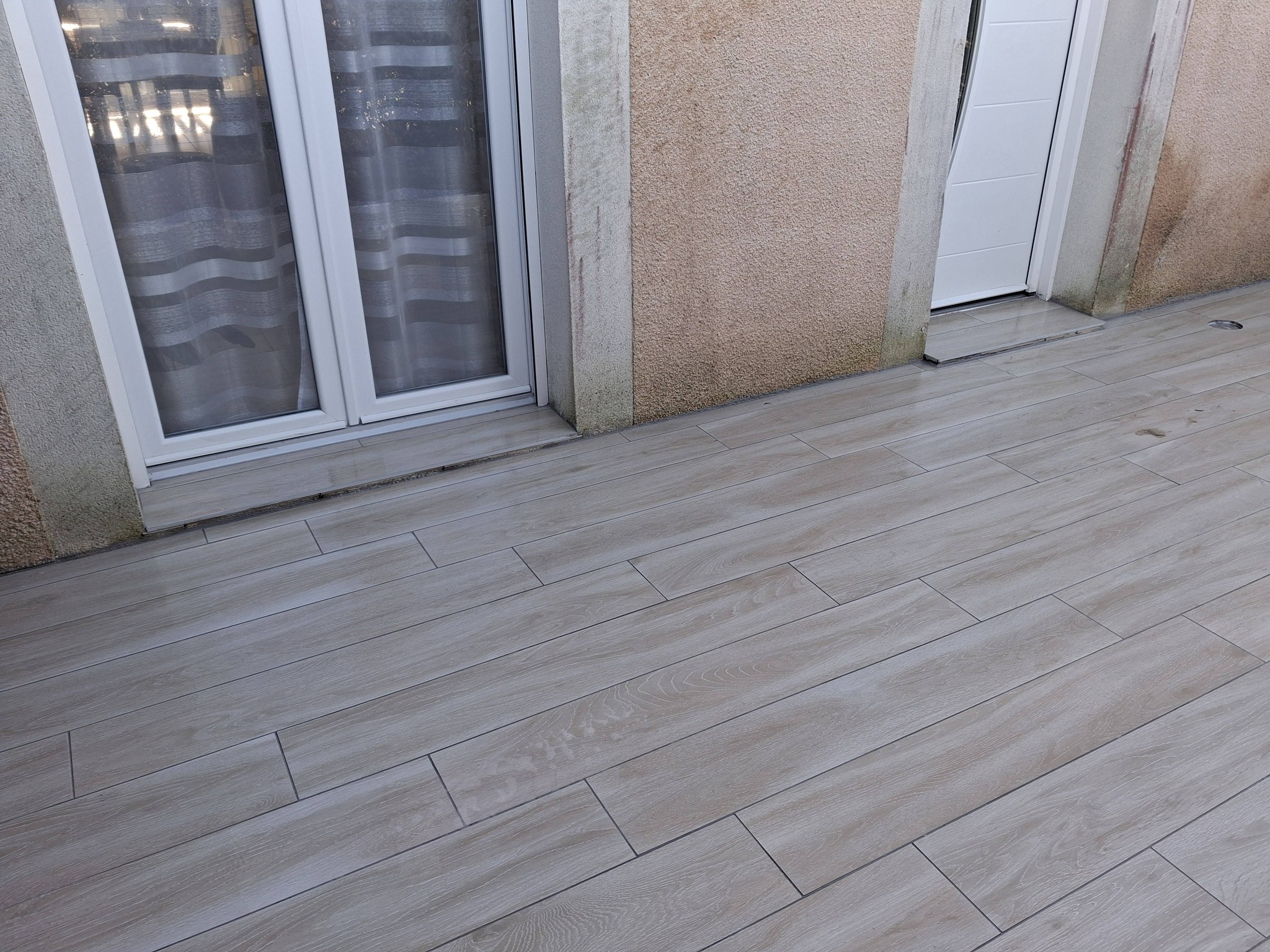 Terrasse extérieure avec carrelage imitation parquet gris clair devant une porte-fenêtre blanche et mur en crépi beige.