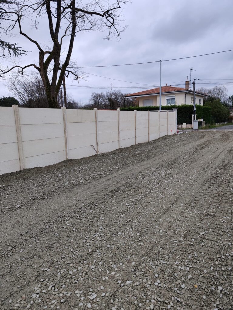 Clôture en béton blanc et allée de gravier pour jardin Clôture en panneaux de béton blanc bordant un grand terrain de gravier devant une maison individuelle.
