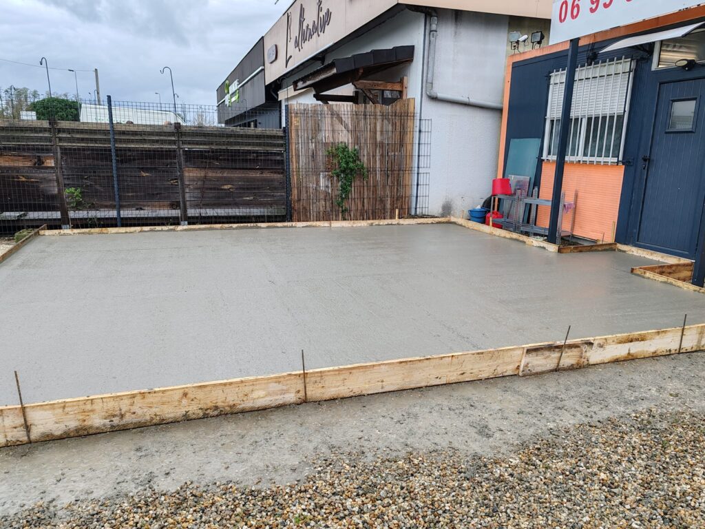 Dalle béton extérieur : coffrage et coulage d'une terrasse Dalle en béton frais récemment coulée avec coffrage en bois devant un bâtiment commercial en extérieur.