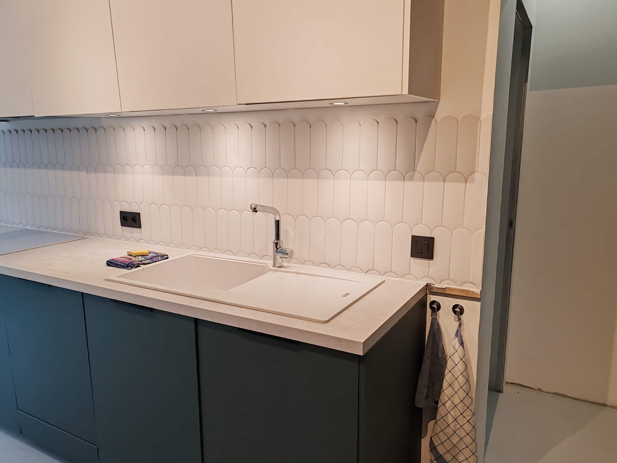 Cuisine moderne avec évier blanc, carrelage en écailles et meubles bas bleu canard sous des placards blancs.