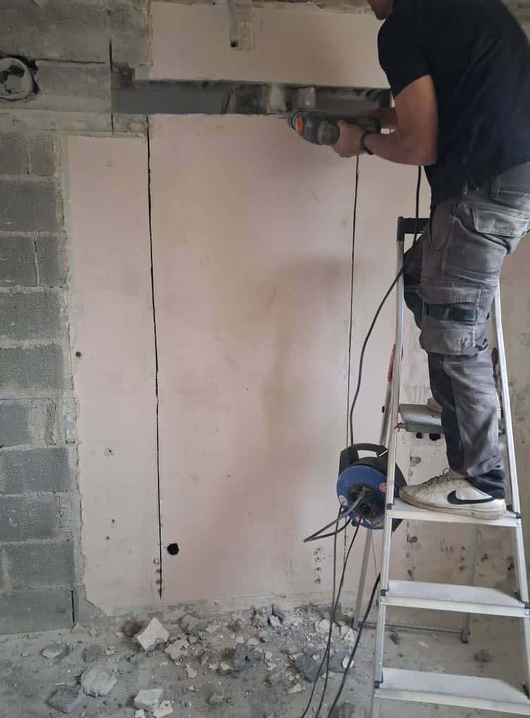 Ouvrier sur un escabeau démolissant un mur intérieur avec un perforateur dans une pièce en rénovation.