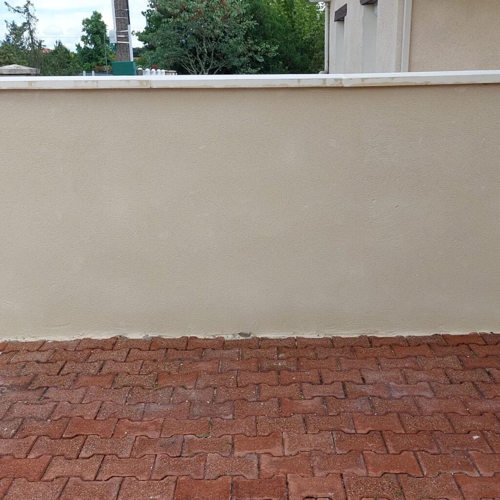 Mur de Clôture Beige et Sol en Pavés Autobloquants Rouges Mur de clôture beige crépi avec chaperon blanc bordant une terrasse en pavés autobloquants rouges en forme de I.