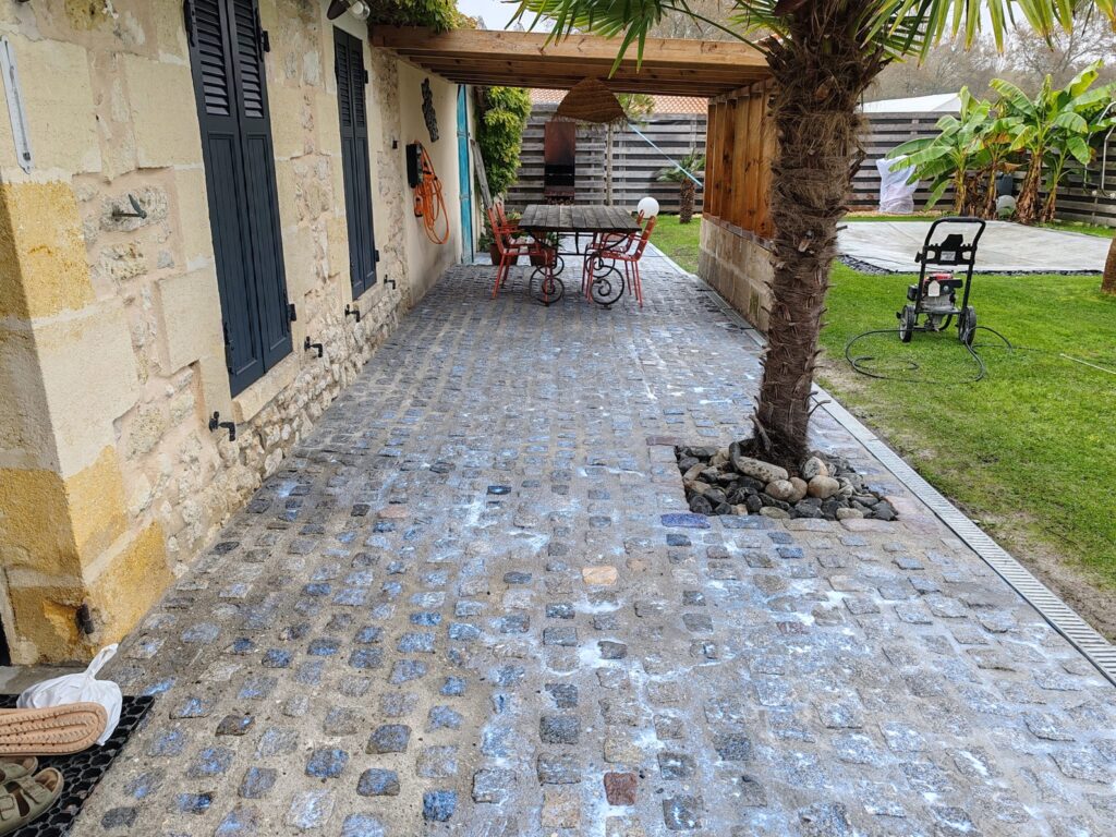 Nettoyage de Patio en Pavés Devant Maison de Pierre Patio en pavés anciens en cours de nettoyage avec mousse devant une maison en pierre et un palmier.