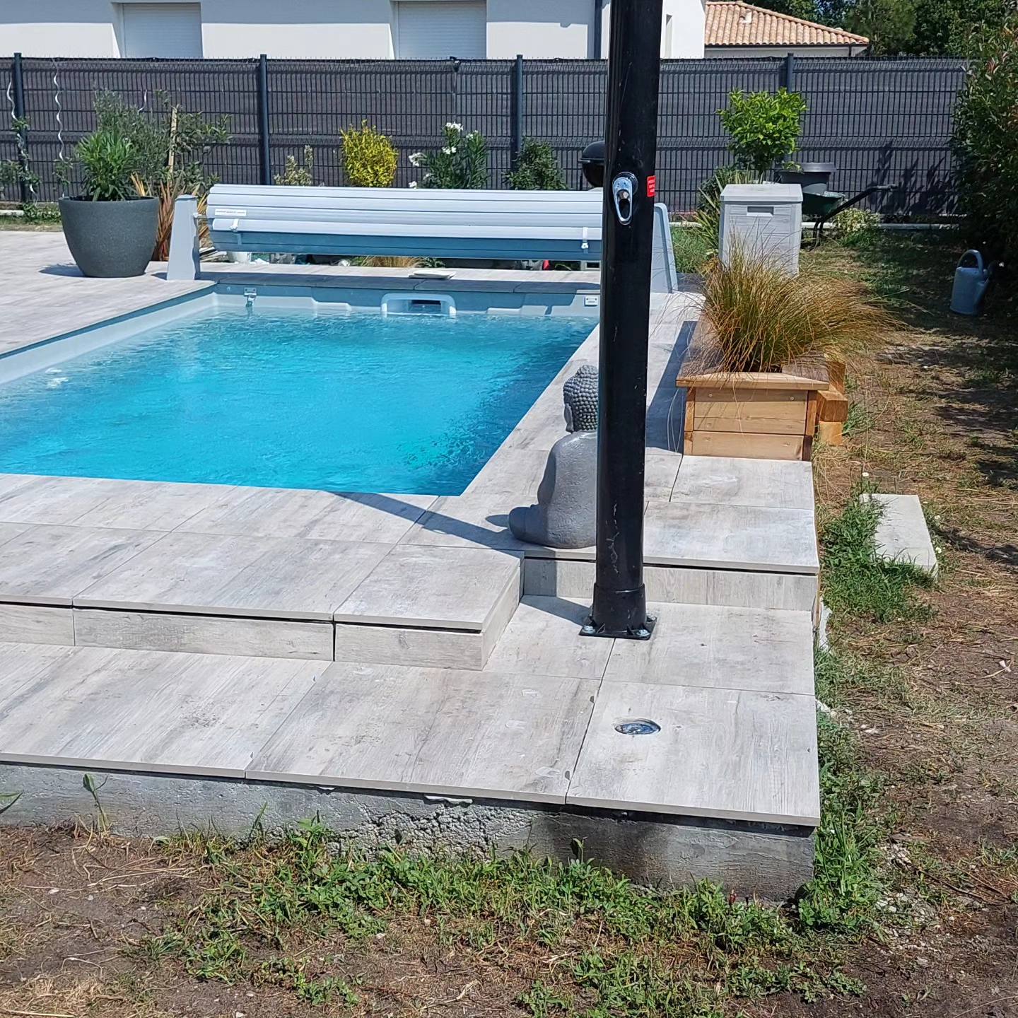 Piscine rectangulaire avec plage en dalles effet bois gris, statue de bouddha, douche noire et bâche de protection.