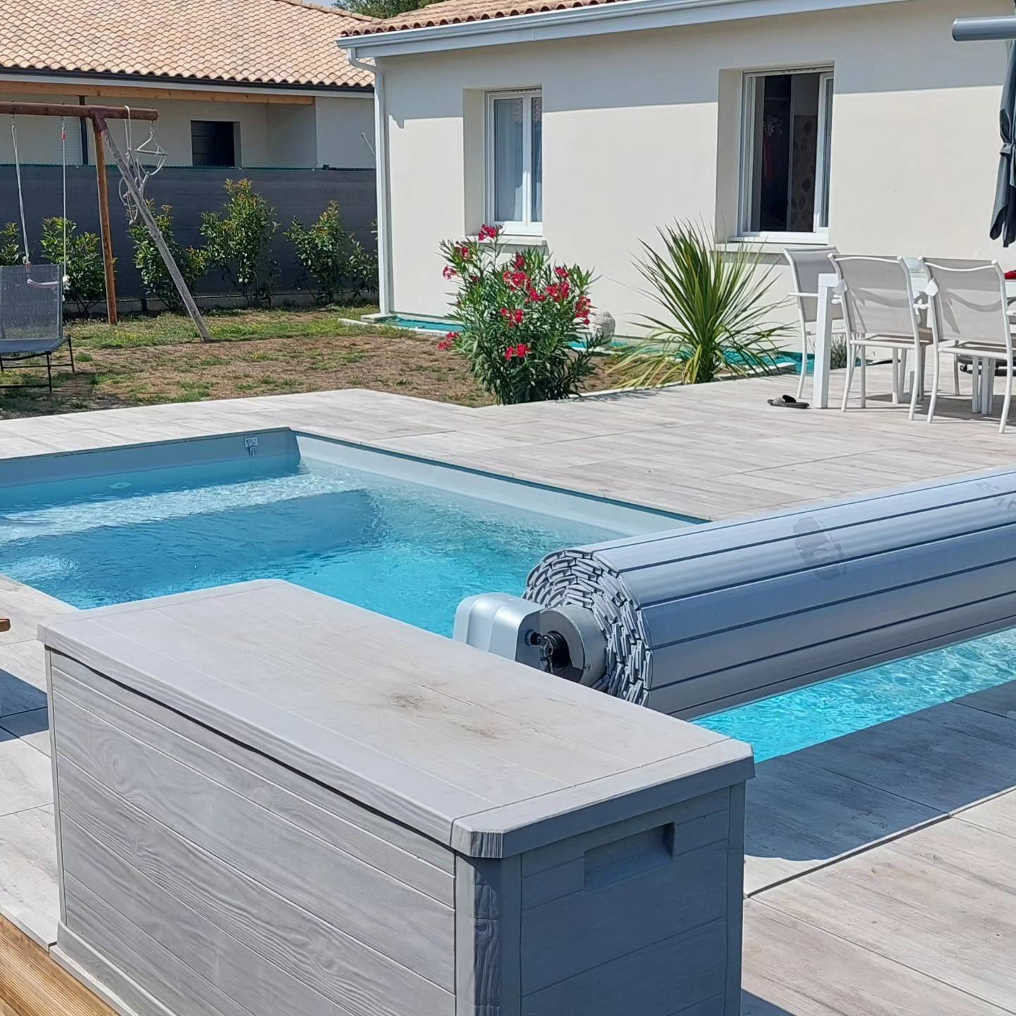 Piscine avec volet roulant sur une terrasse en carrelage aspect bois, jardin aménagé et maison moderne.