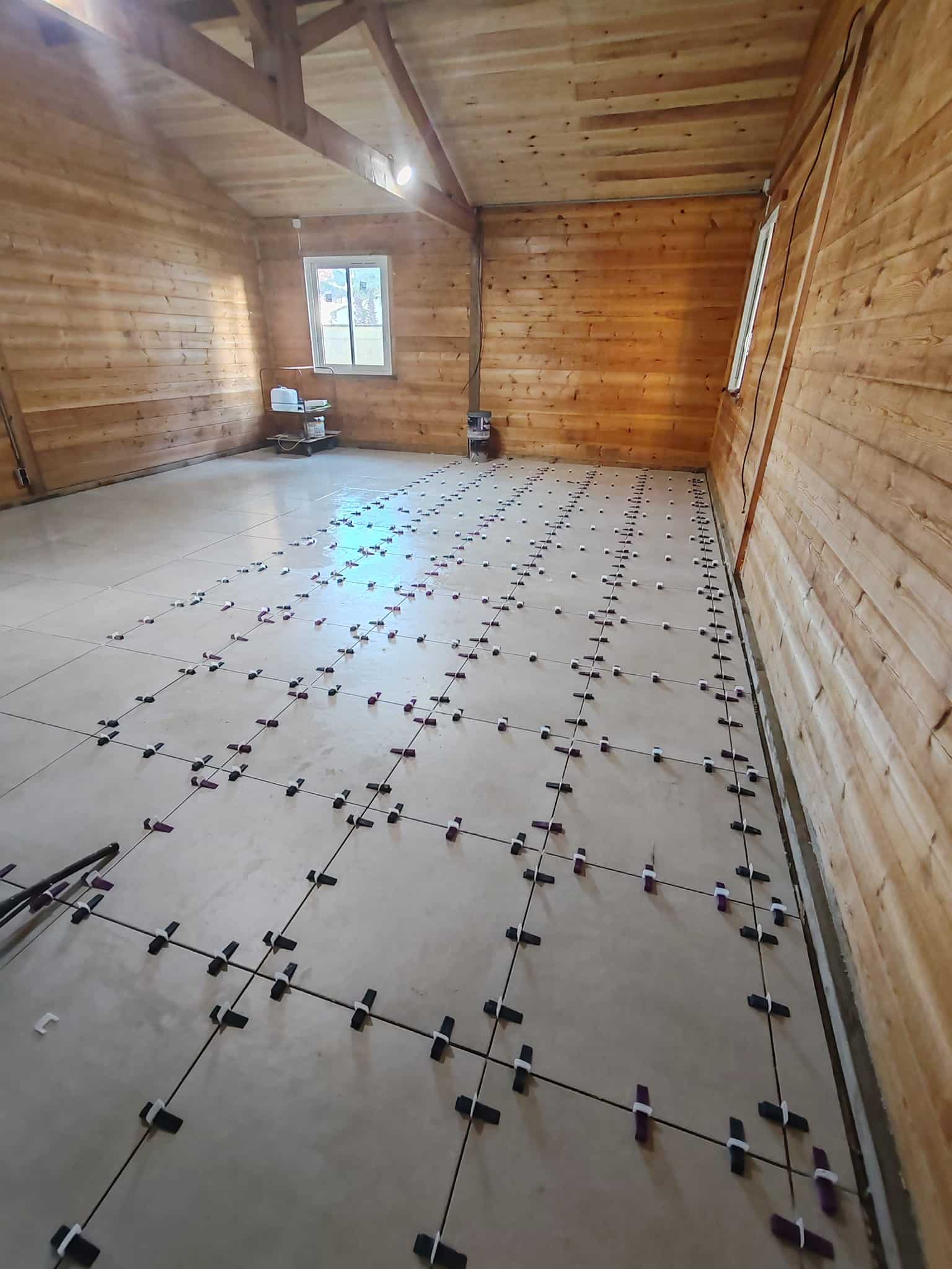 Pose de carrelage grand format avec croisillons autonivelants dans une pièce aux murs en bois rustiques.