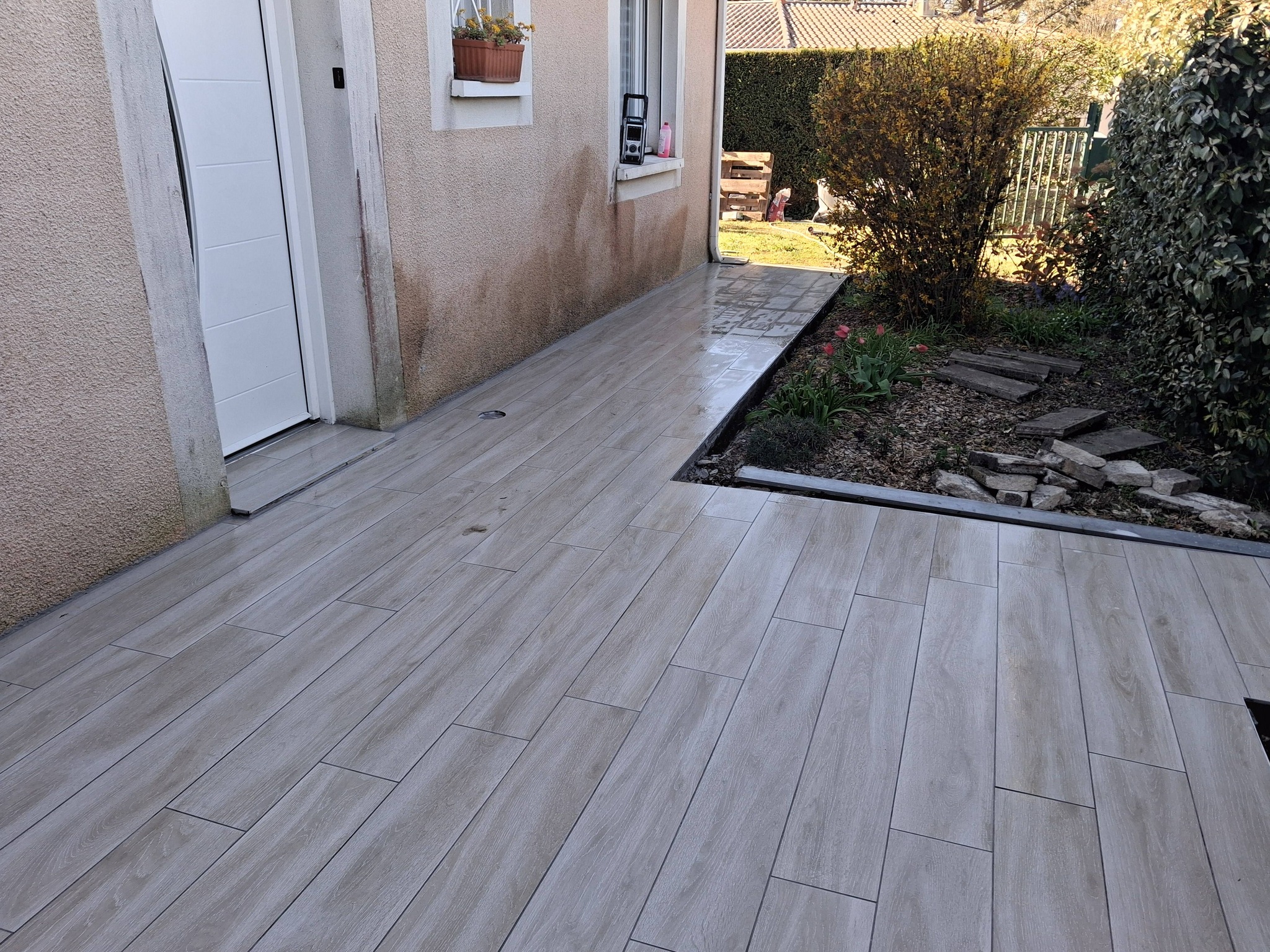 Terrasse moderne en carrelage gris effet bois longeant une maison avec jardin paysager en arrière-plan.