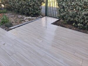 Terrasse moderne en carrelage imitation bois gris clair bordée de haies de jardin menant à un portillon.