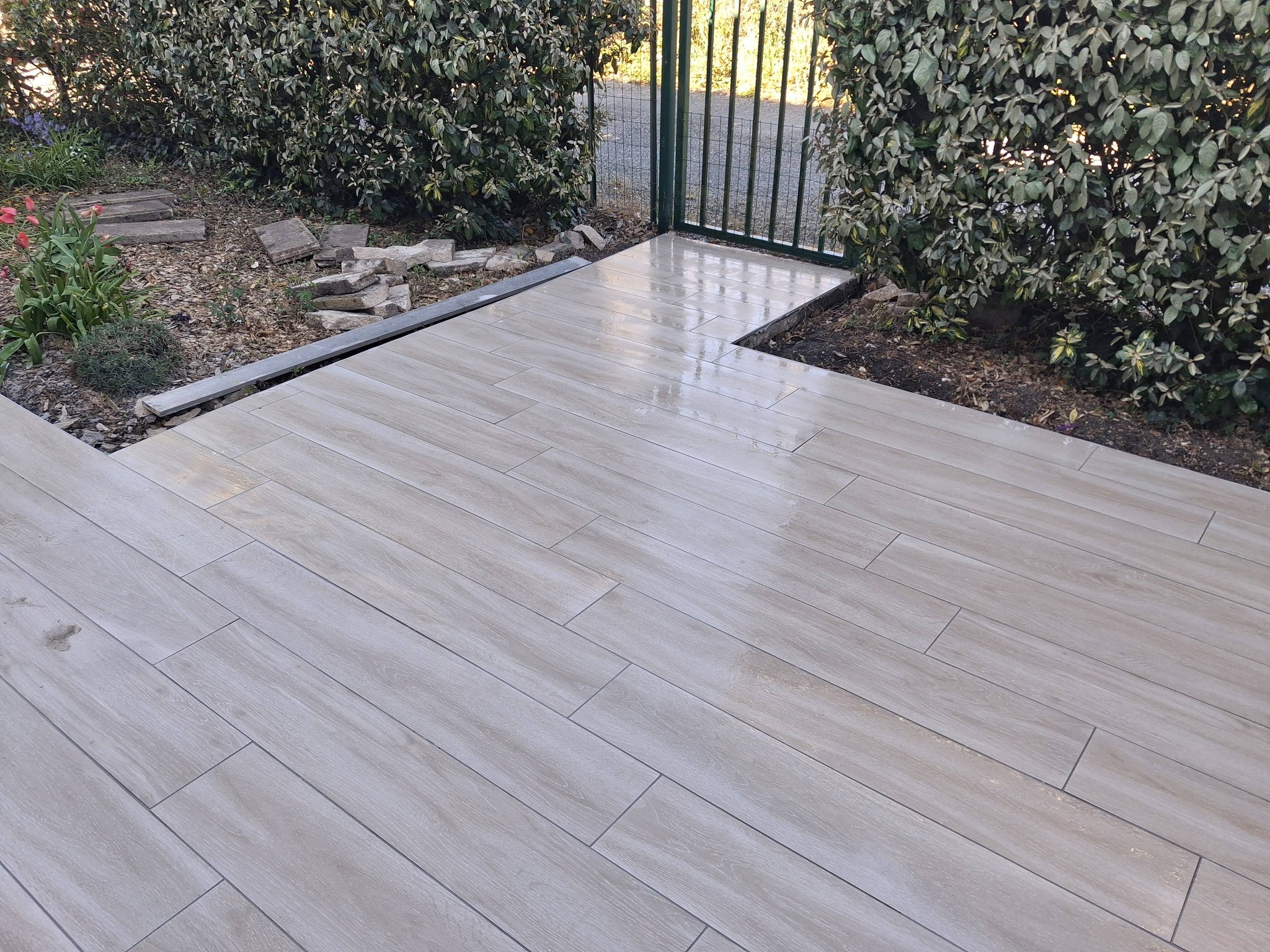Terrasse moderne en carrelage imitation bois gris clair bordée de haies de jardin menant à un portillon.
