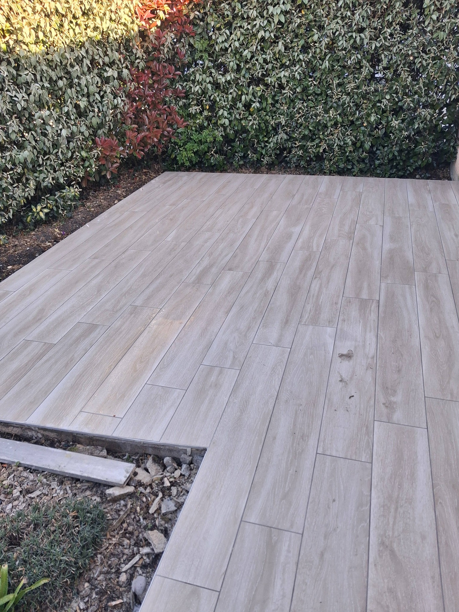Terrasse en carrelage imitation bois clair bordée par une haie de jardin dense et verdoyante.