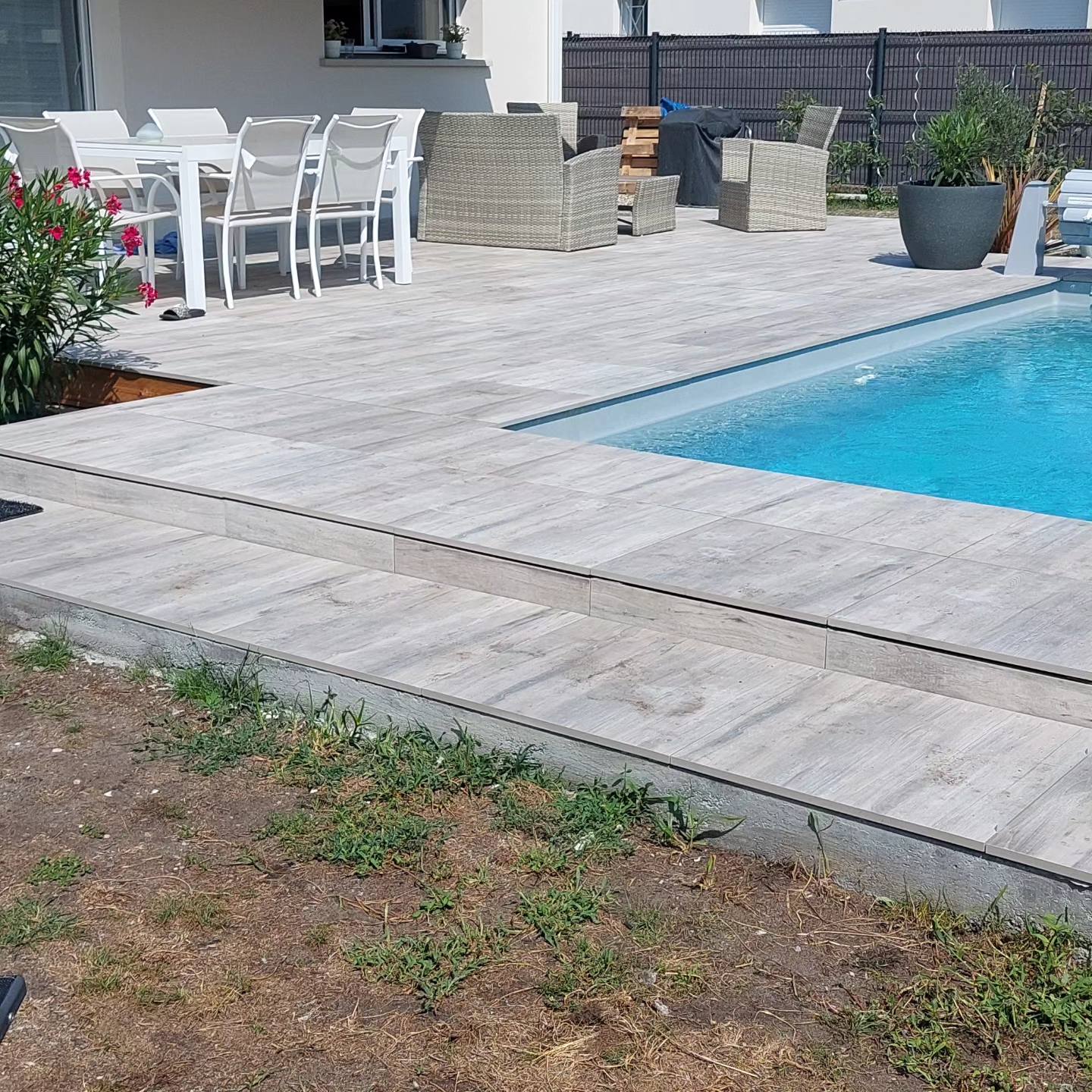 Terrasse moderne en carrelage aspect bois gris clair bordant une piscine avec mobilier de jardin blanc et osier.
