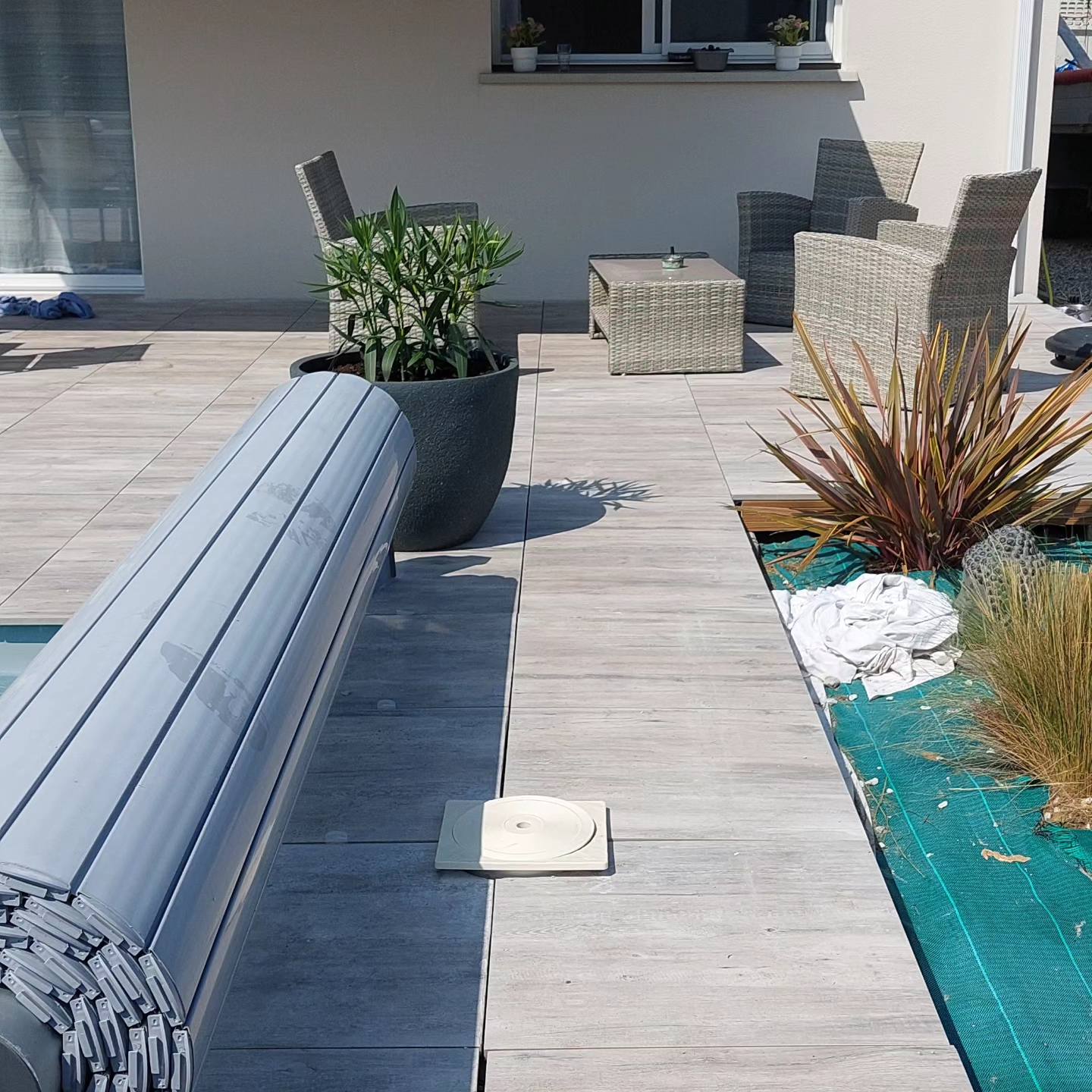 Volet roulant de piscine gris sur une terrasse en carrelage avec salon de jardin en rotin et plantes décoratives.